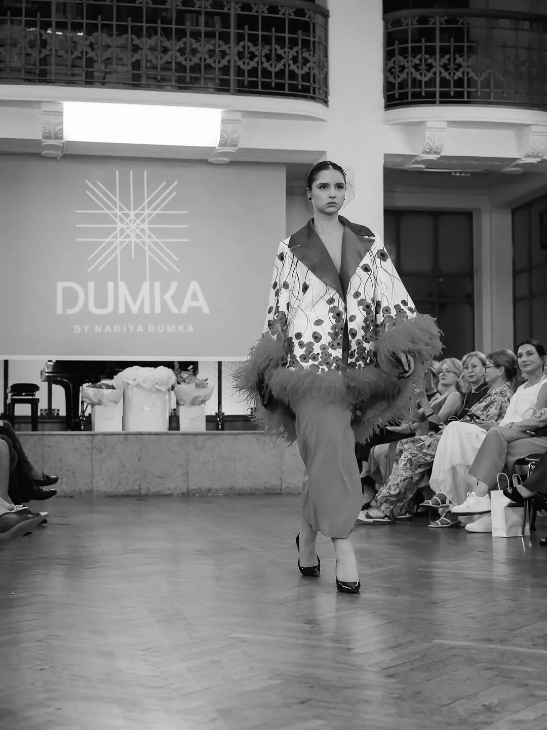 Sort-hvidt foto af model i frakke med fjer på catwalken foran DUMKA-skiltet